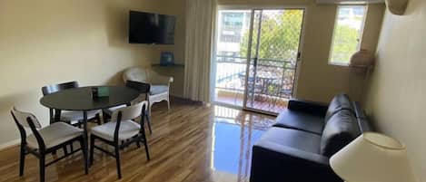 A room with a dining table and chairs, a sofa, a TV mounted on the wall, and a window with a view of the outside.
