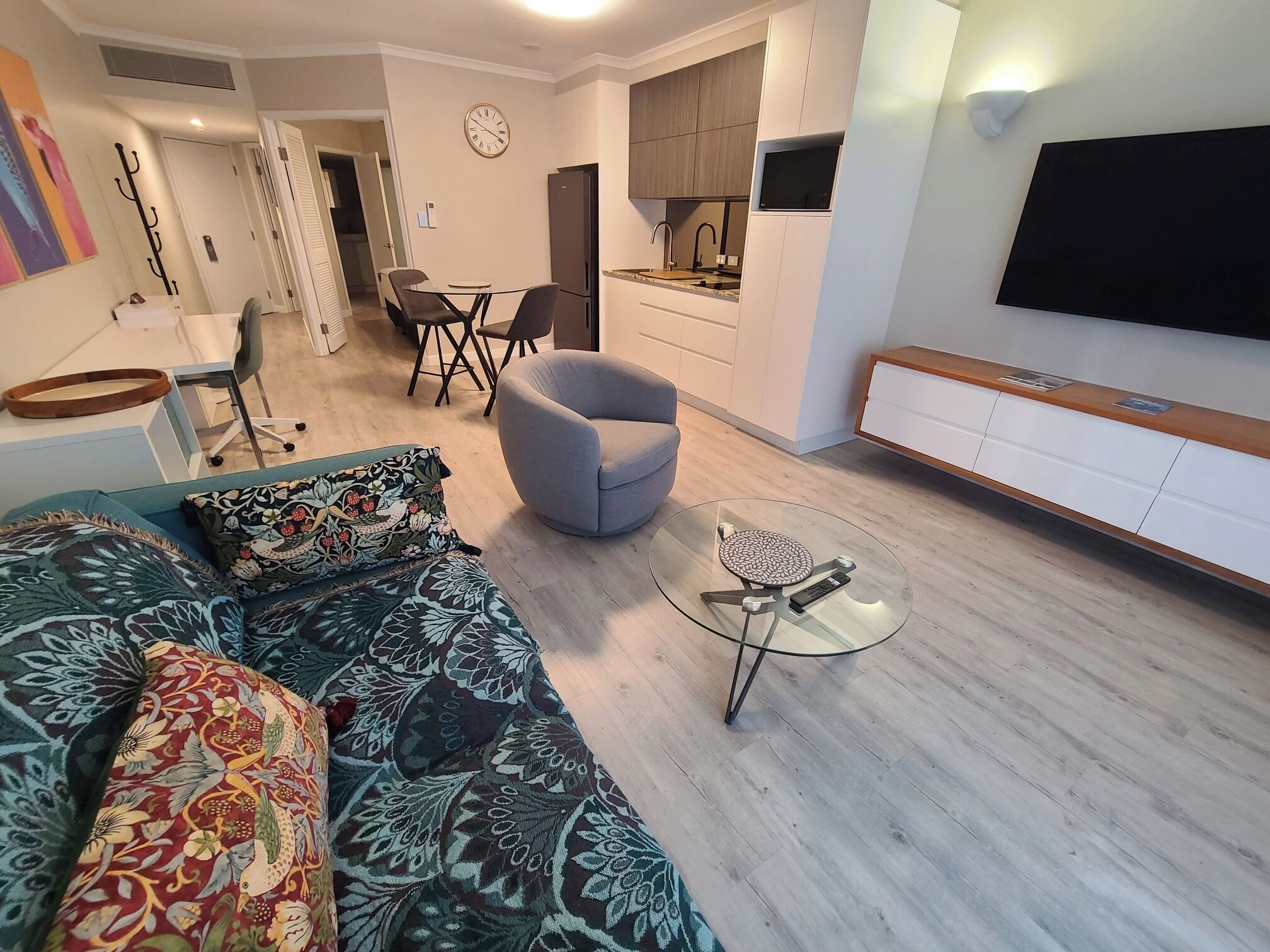 A modern living room with a sofa, armchair, and coffee table. There is a dining area with chairs and a table, and a kitchen with cabinets and a TV mounted on the wall.