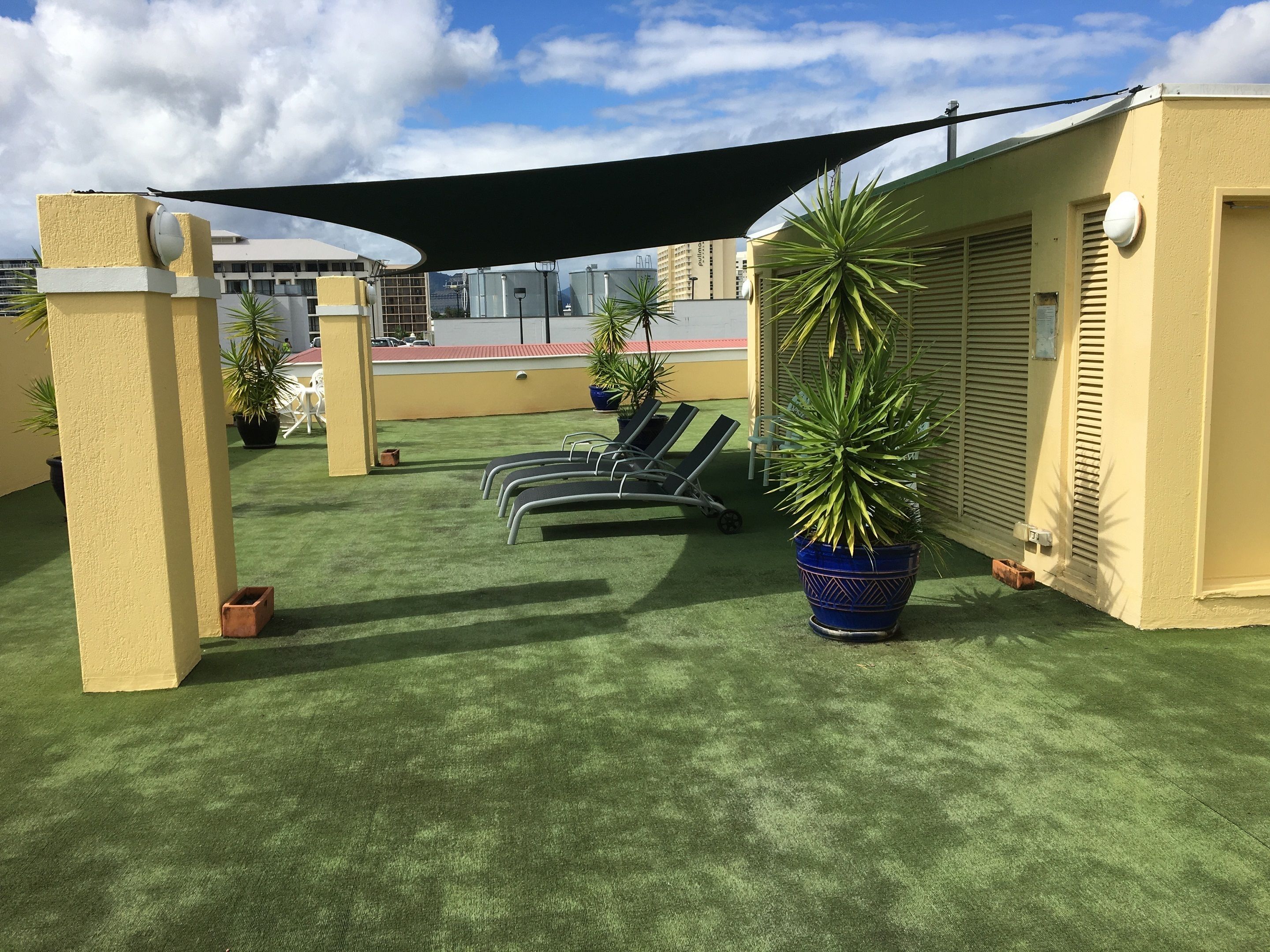 A rooftop terrace with artificial grass, lounge chairs, potted plants, and a large shade sail.