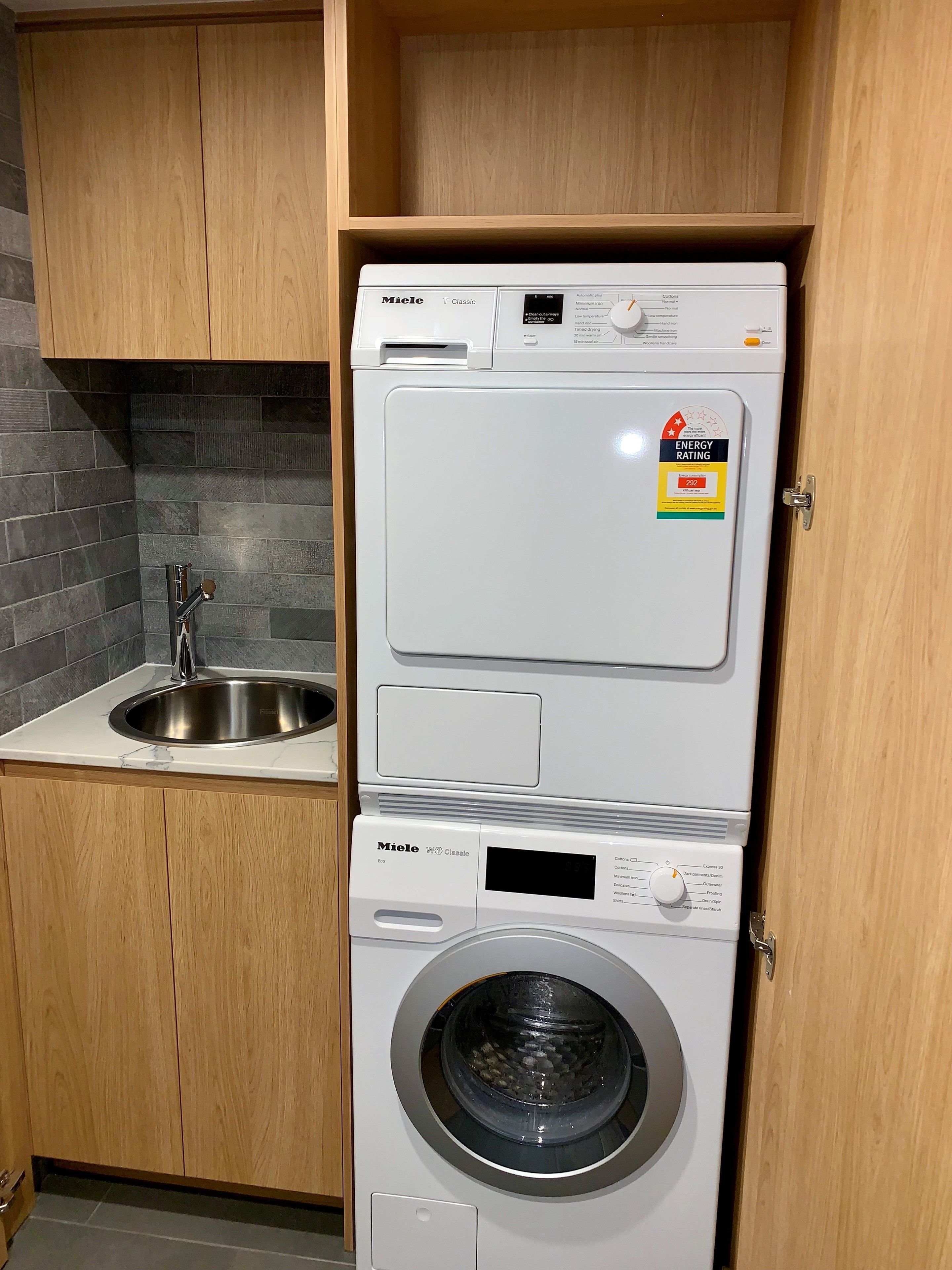 A compact, two-door laundry machine with a digital display and energy rating sticker, situated in a kitchen with a sink and wooden cabinets.