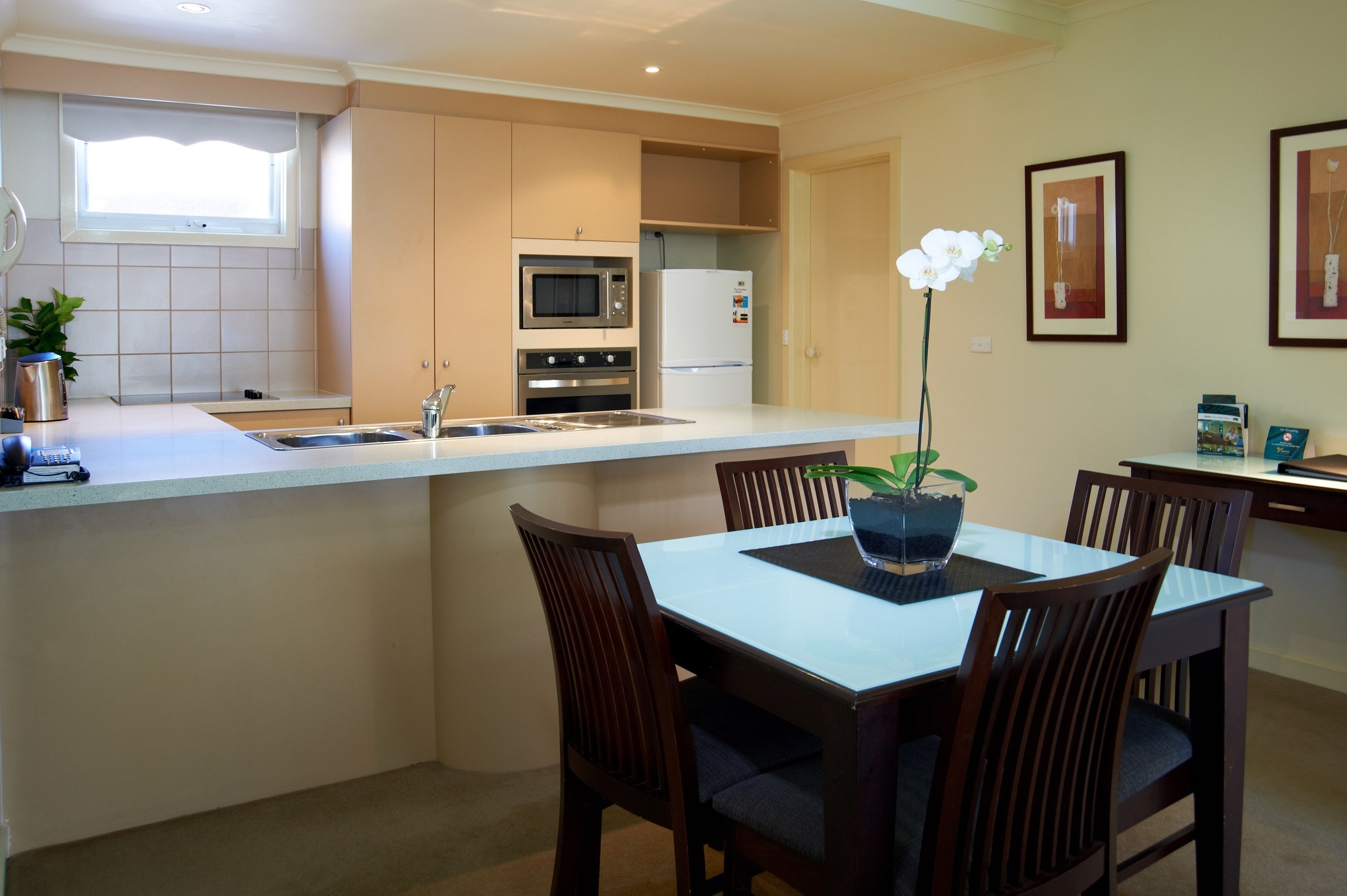 Une cuisine moderne dotée d’un îlot central, d’un four encastré, d’un micro-ondes et d’un réfrigérateur. On y trouve également un coin repas avec une table et des chaises, ainsi qu’une orchidée en pot sur l’îlot.
