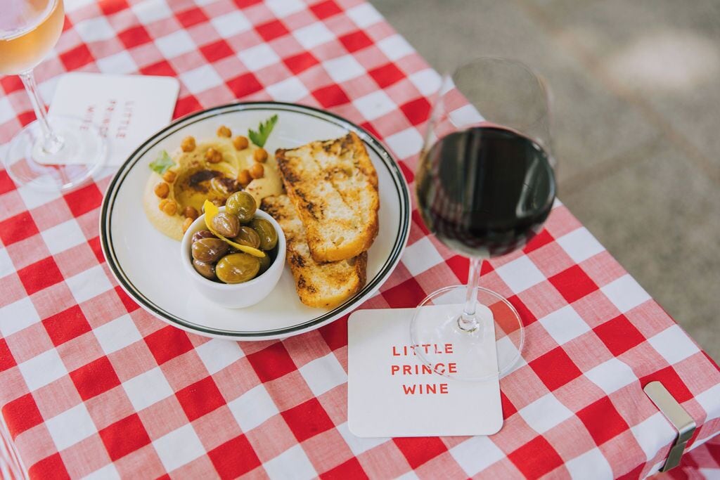 A meal with a fish, olives, and a small bowl of sauce, served on a checkered tablecloth with a wine glass and a tag reading "Little Prince Wine".