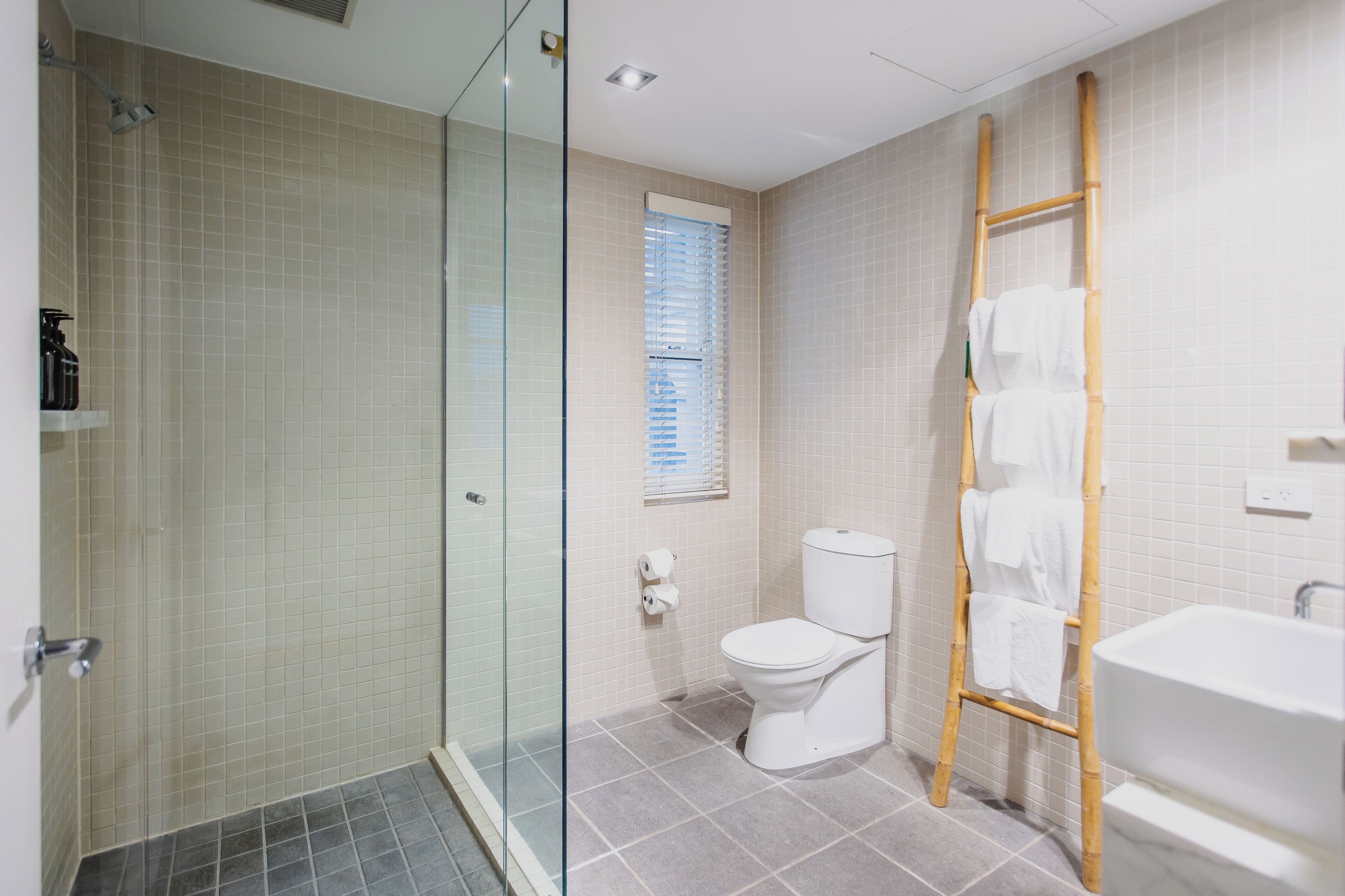 Une salle de bain avec une douche à parois de verre, des toilettes, une fenêtre avec des stores et une échelle en bois sur laquelle sont posées des serviettes.