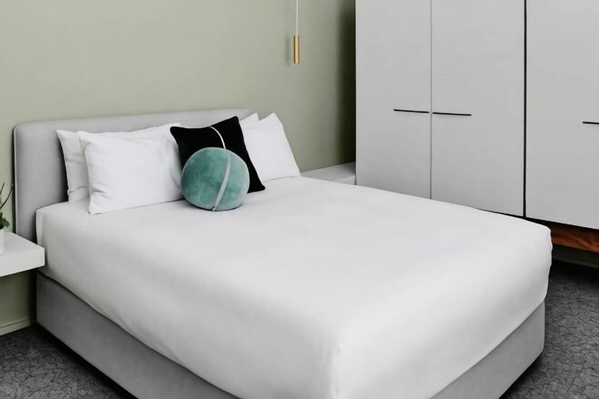 A neatly made bed with white bedding and a teal pillow, a white nightstand with a plant, and a white wardrobe with horizontal handles.