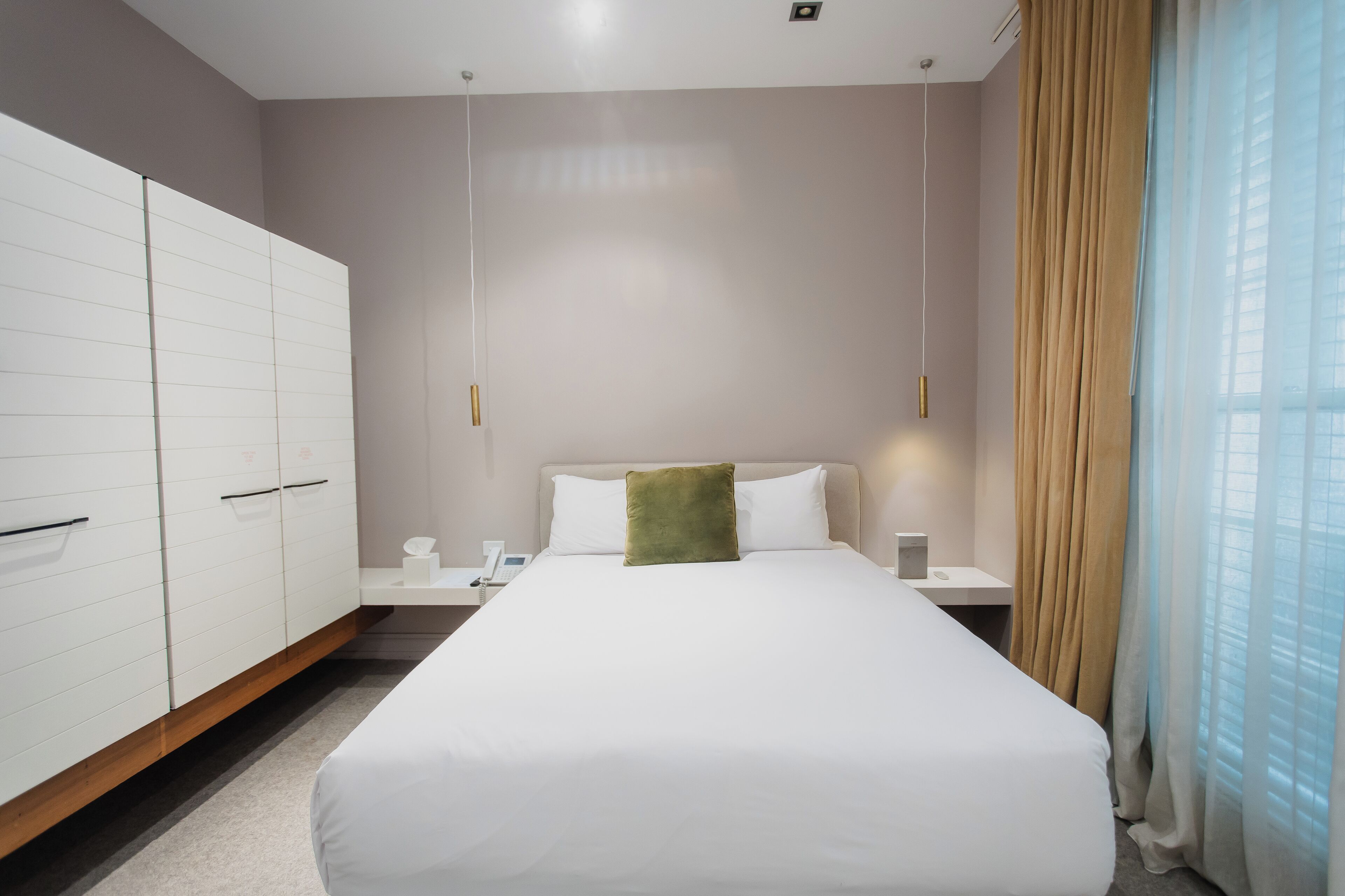 Una habitación de hotel moderna con una cama grande, un armario blanco, mesitas de noche y vistas de la ciudad a través de cortinas translúcidas.