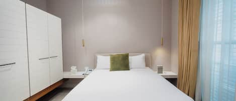 A modern hotel room with a large bed, white wardrobe, bedside tables, and a view of the city through sheer curtains.