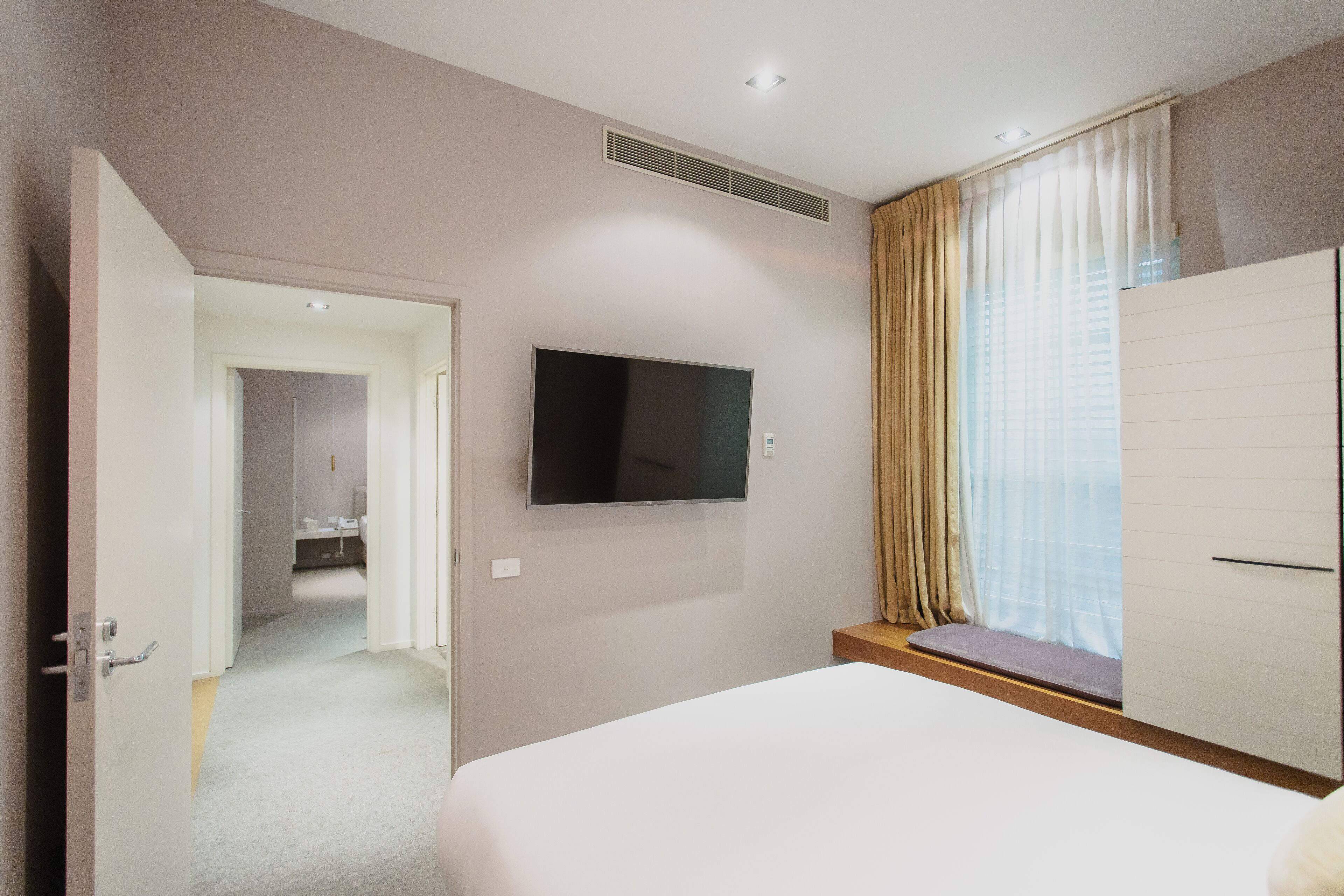 A hotel room with a bed, a TV mounted on the wall, a window with curtains, and a door leading to another room.