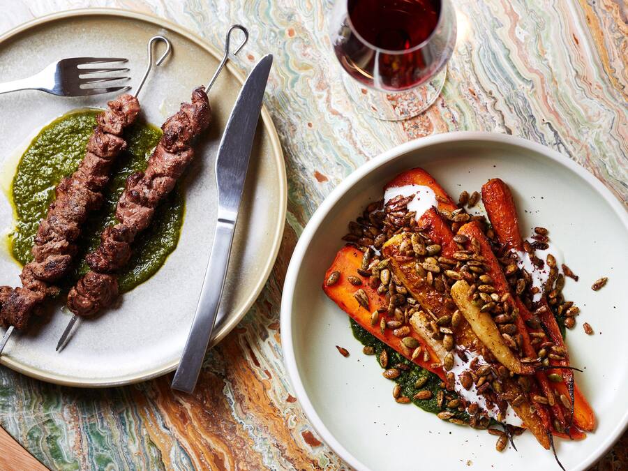 En tallerken med spydstegt kød med grøn sauce, en tallerken med ristede grøntsager med nødder, og et glas rødvin på et marmorbord.