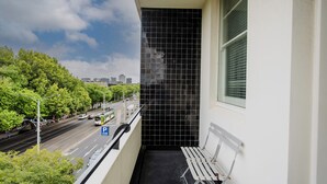 A balcony with a black tiled wall, a white bench, and a view of a city street with cars and greenery.