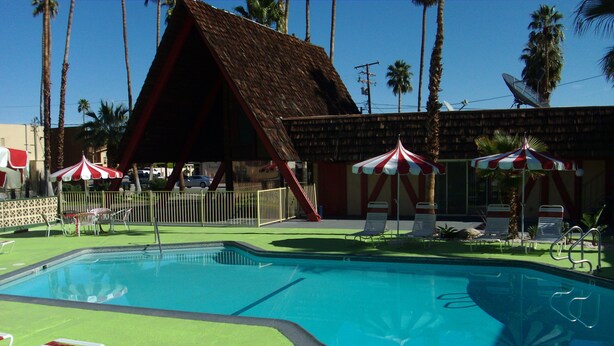 A swimming pool with lounge chairs and umbrellas, surrounded by palm trees and a building with a thatched roof. - Desert Lodge (Palm Springs)