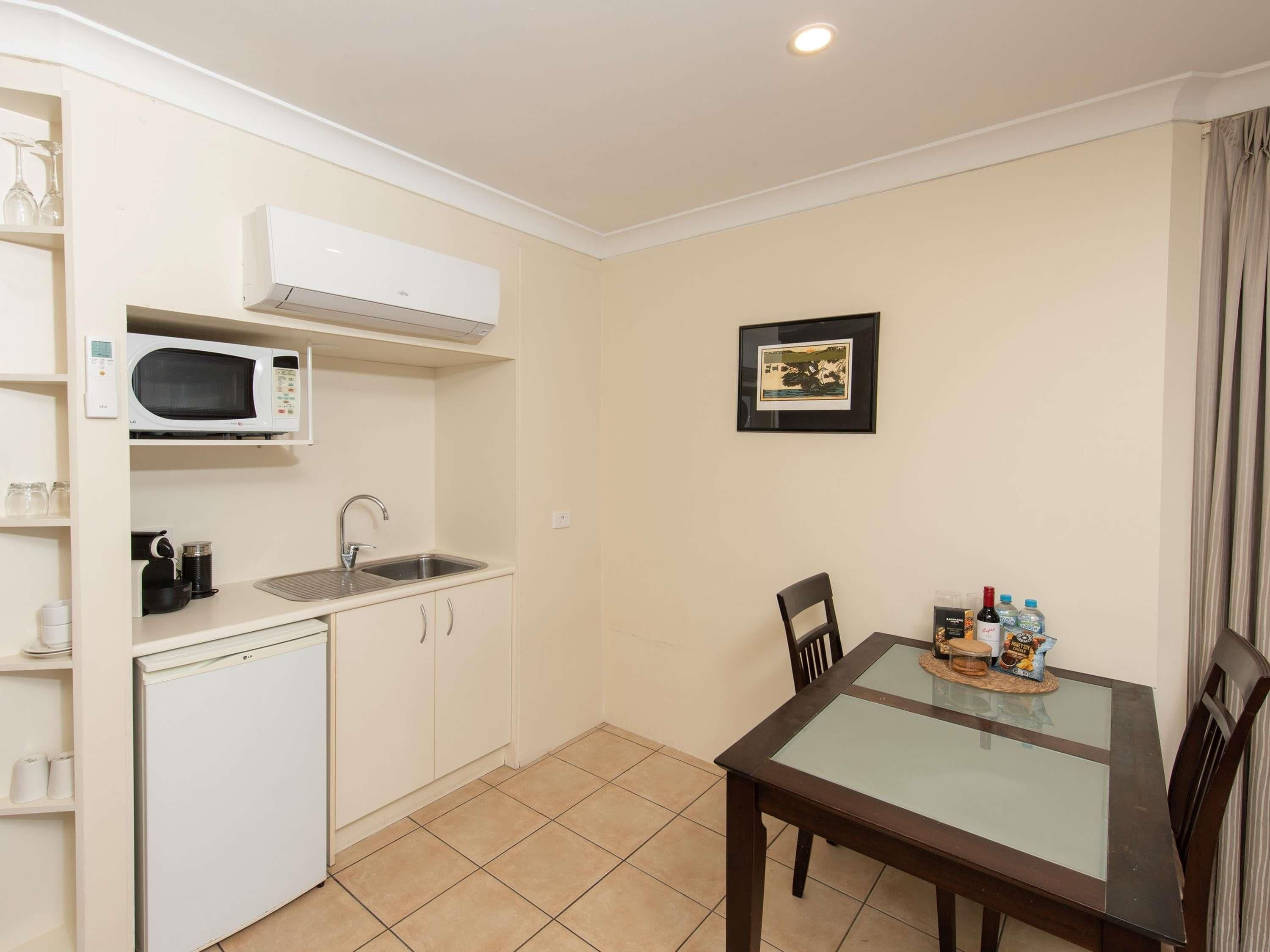 A compact kitchen with a microwave, sink, and cabinets, a dining table with chairs, and a framed picture on the wall.
