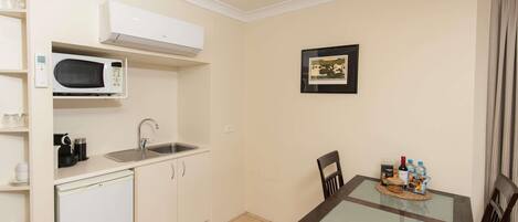 A compact kitchen with a microwave, sink, and cabinets, a dining table with chairs, and a framed picture on the wall.