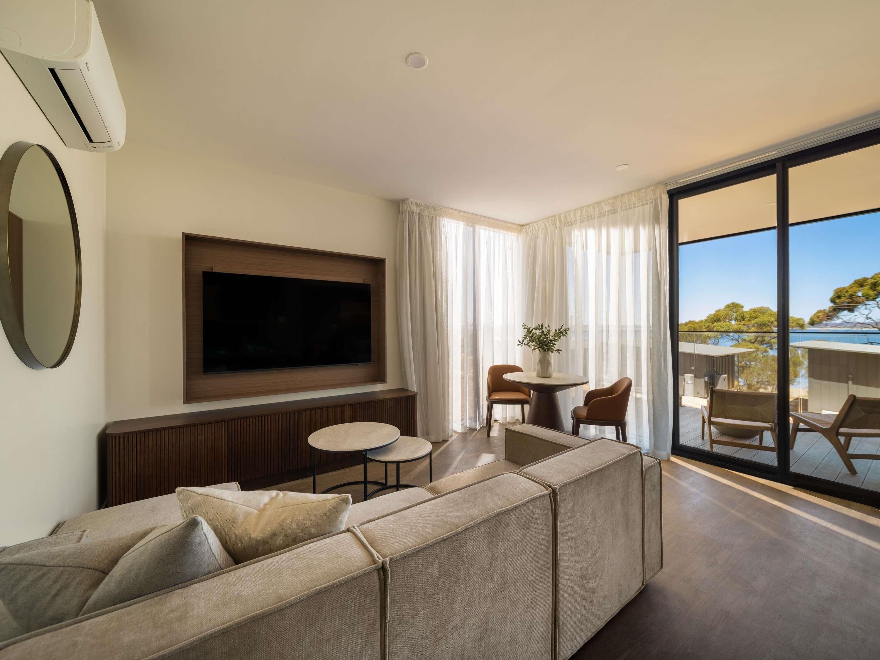 A modern living room with a large flat-screen TV, a sectional sofa, a small round table with chairs, and a balcony with a view.