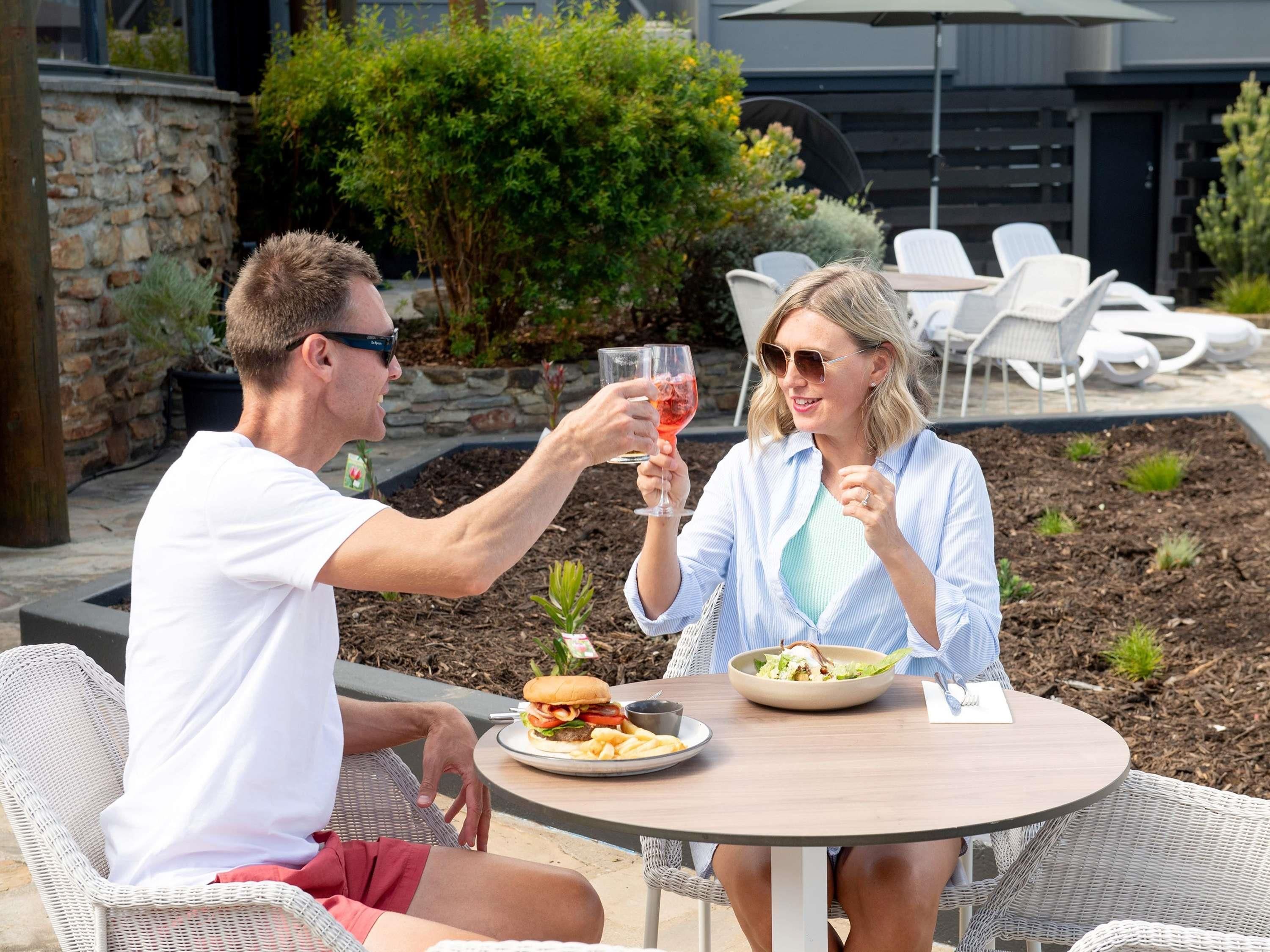 Two people seated at an outdoor table with a meal, surrounded by outdoor furniture and greenery.