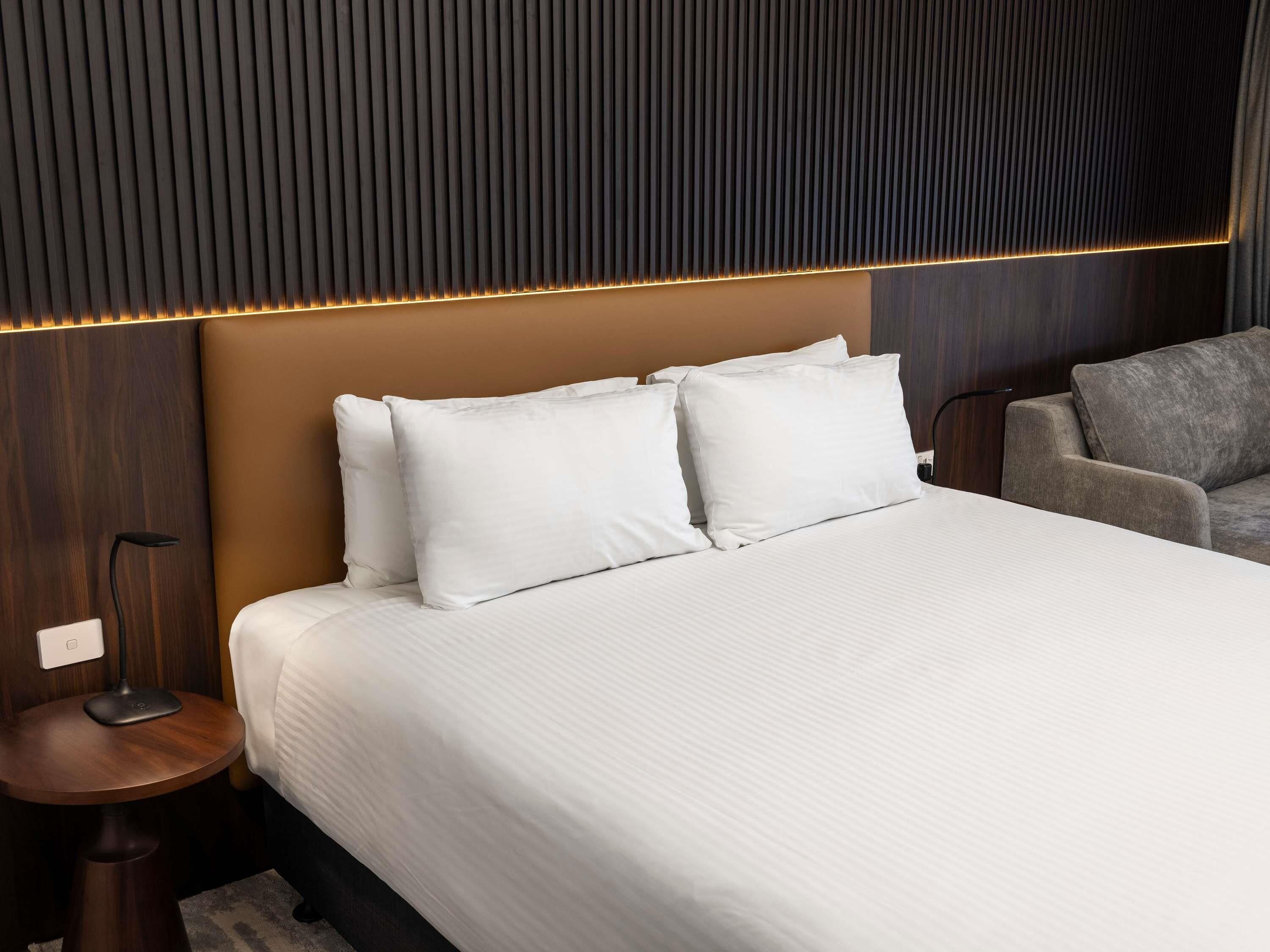Una habitación de hotel con una cama grande, dos almohadas, una mesita de noche con una lámpara y un sillón gris.
