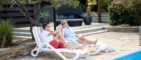 Two people relaxing on white lounge chairs by a pool with a stone patio and a black outdoor sofa in the background.