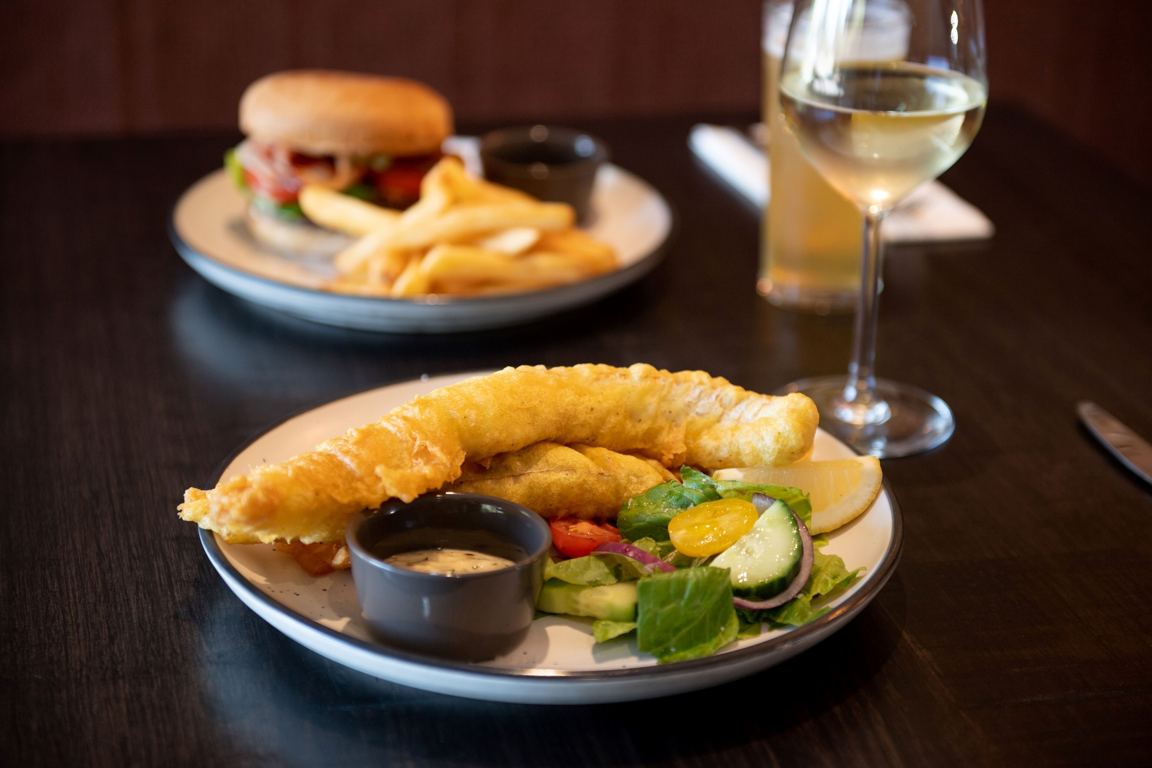 Un plato con ensalada, patatas fritas y un sándwich, junto con una copa de vino blanco sobre una mesa.