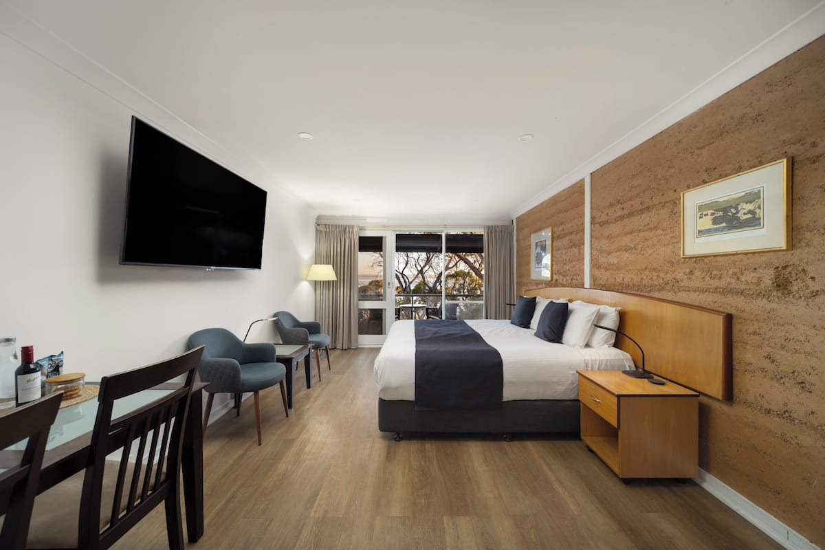 Een moderne hotelkamer met een groot bed, een flatscreen televisie, een zithoek met een tafel en stoelen, en uitzicht door een glazen deur.