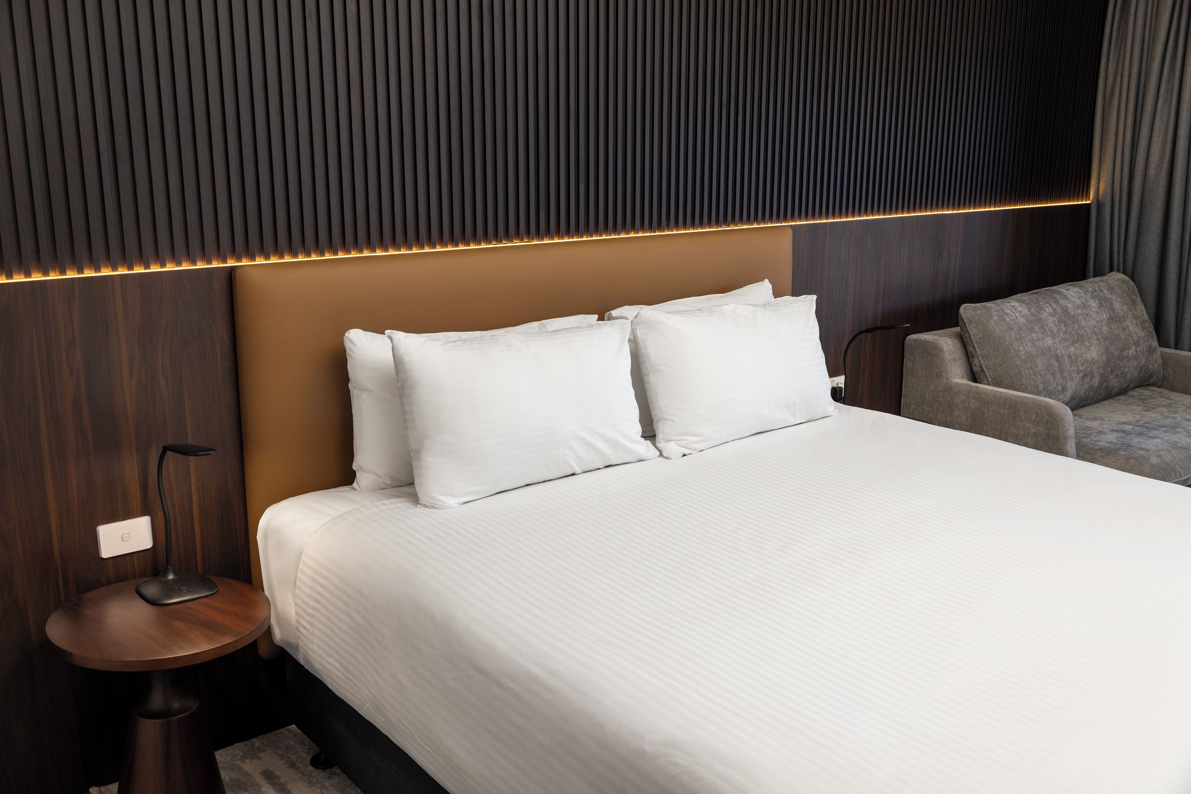 A modern hotel room with a large bed, a bedside table, a lamp, and a grey armchair.