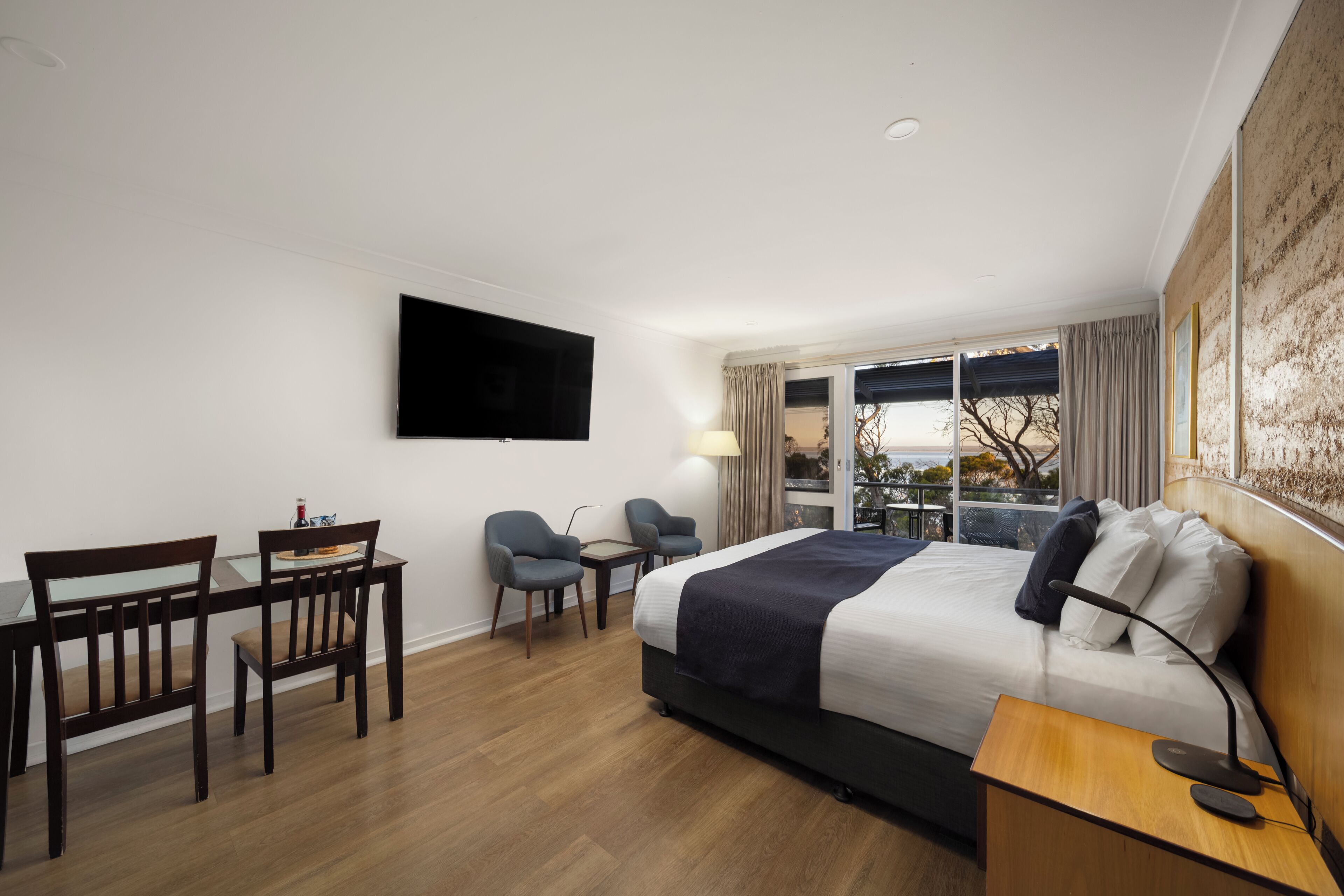 Una habitación de hotel con una cama, un escritorio, dos sillas, un televisor montado en la pared y una ventana con cortinas.