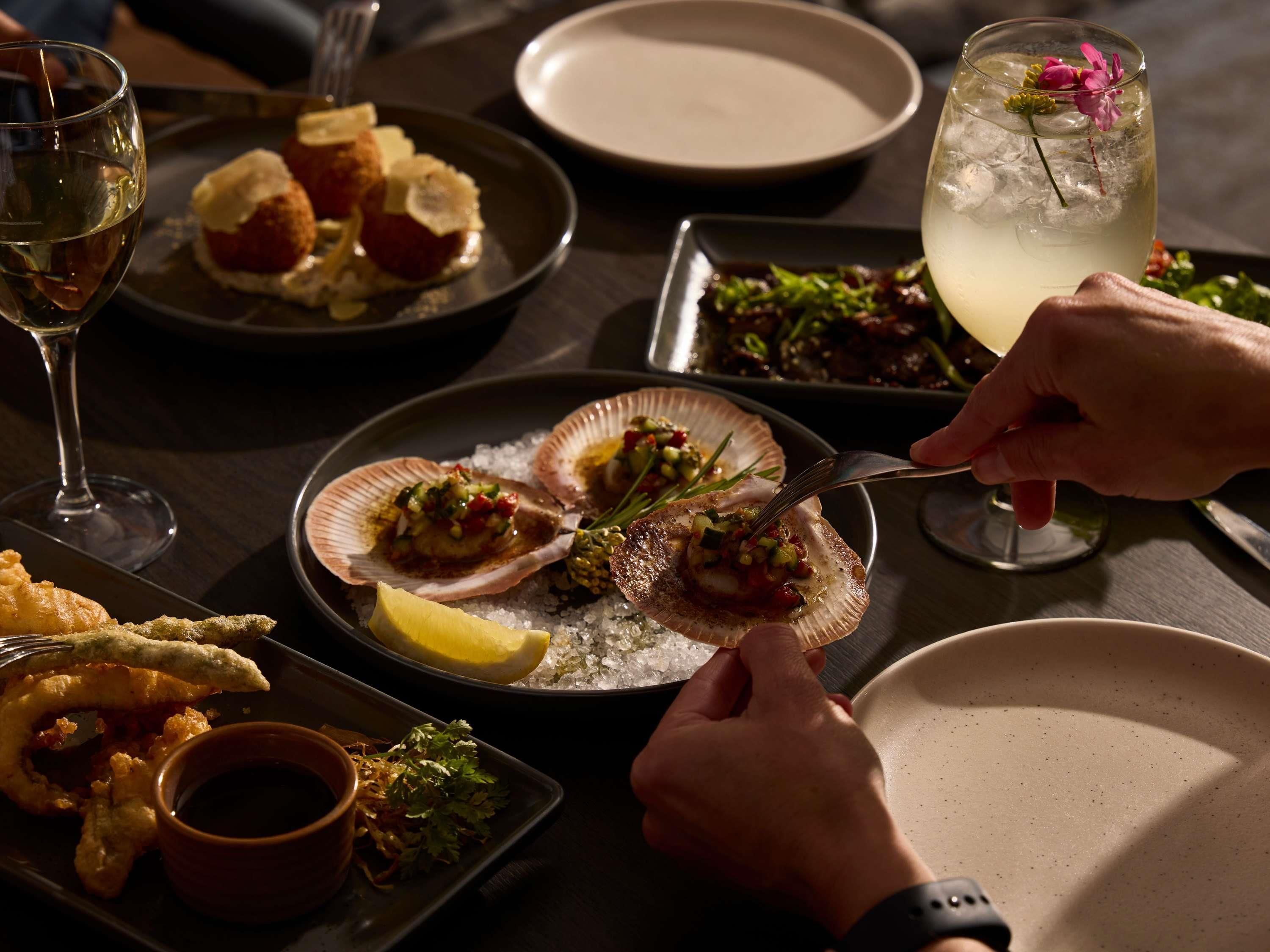 A dining table with seafood dishes, a glass of white wine, and a cocktail with a flower garnish.
