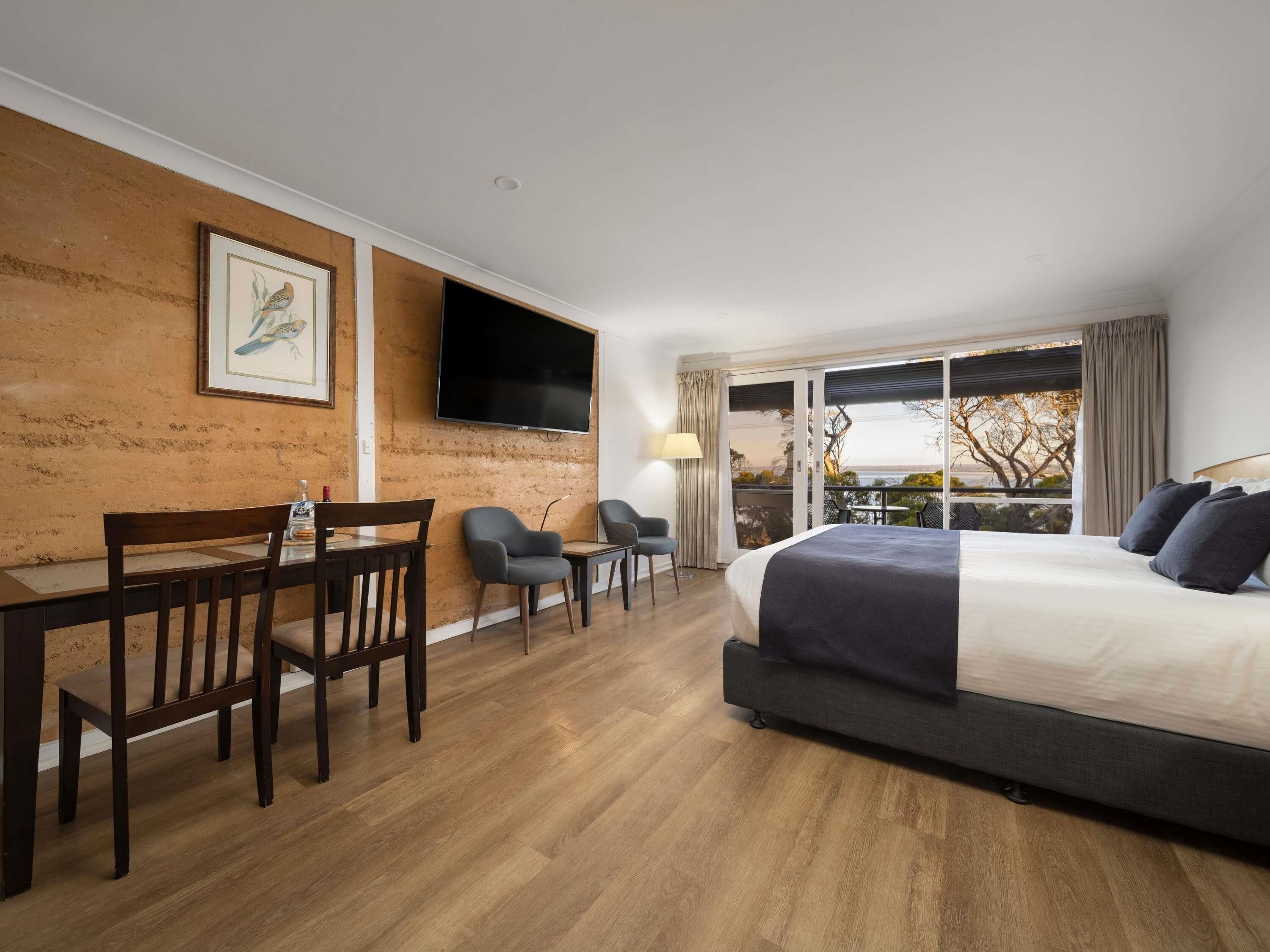 A hotel room with a bed, a desk with chairs, a TV mounted on the wall, and a balcony with a view.