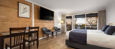 A hotel room with a bed, a desk with chairs, a TV mounted on the wall, and a balcony with a view.