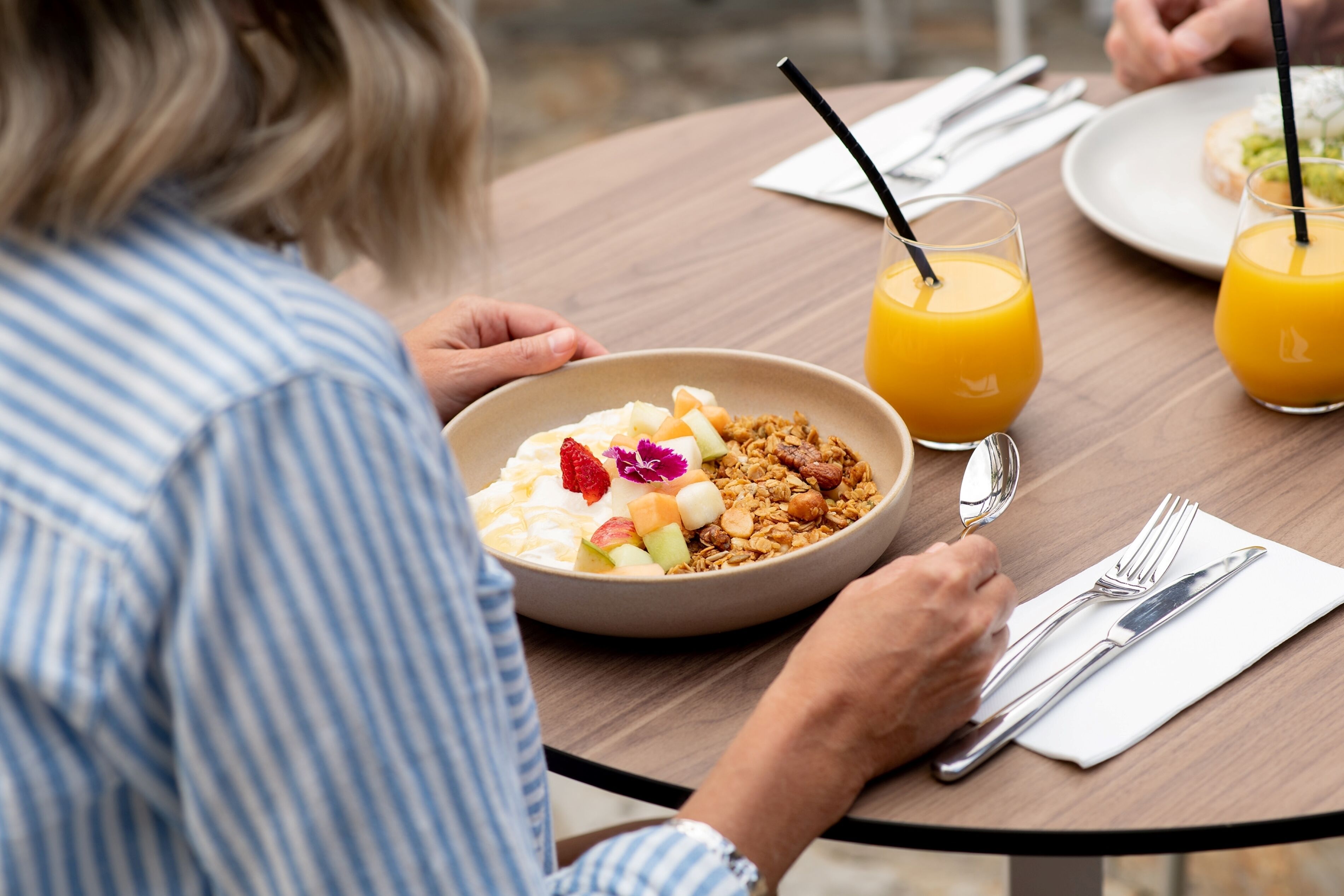 A dining table with a bowl of cereal, two glasses of orange juice, and cutlery.