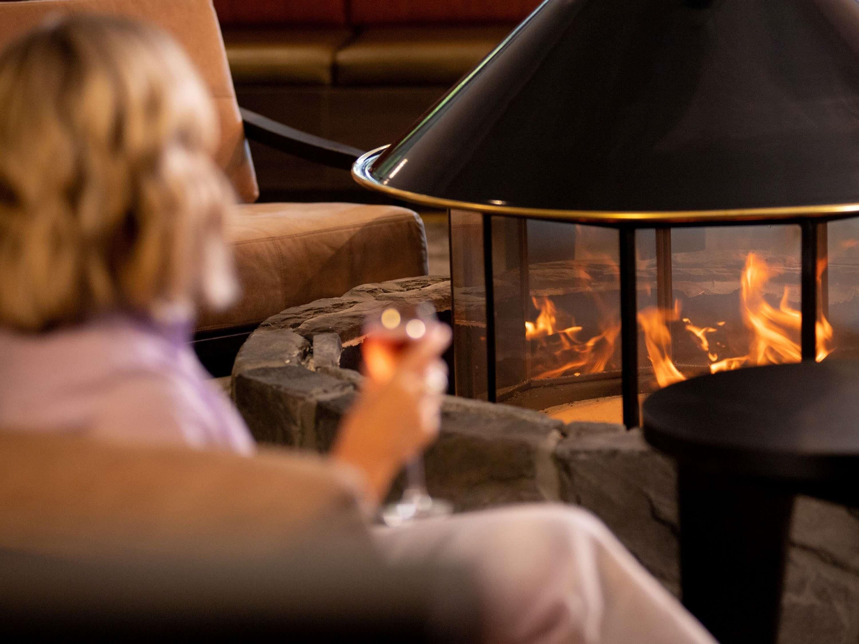 Una persona sentada junto a una chimenea de vidrio con llamas visibles, una mesa de piedra y un sillón acolchado.