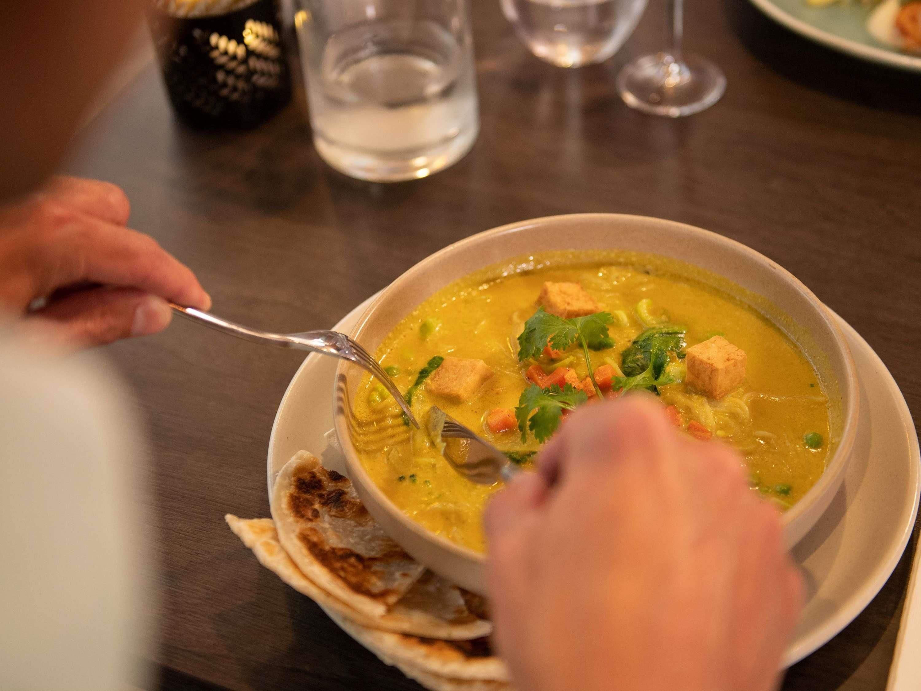 A person eating a bowl of soup with a spoon, with a glass of water and a plate of flatbread on the table.