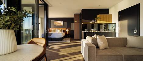 A modern hotel room with a large bed, a sofa, a dining table, and a kitchen area.