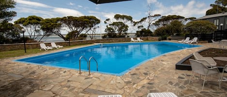 A swimming pool with stone flooring, surrounded by white lounge chairs and an umbrella.