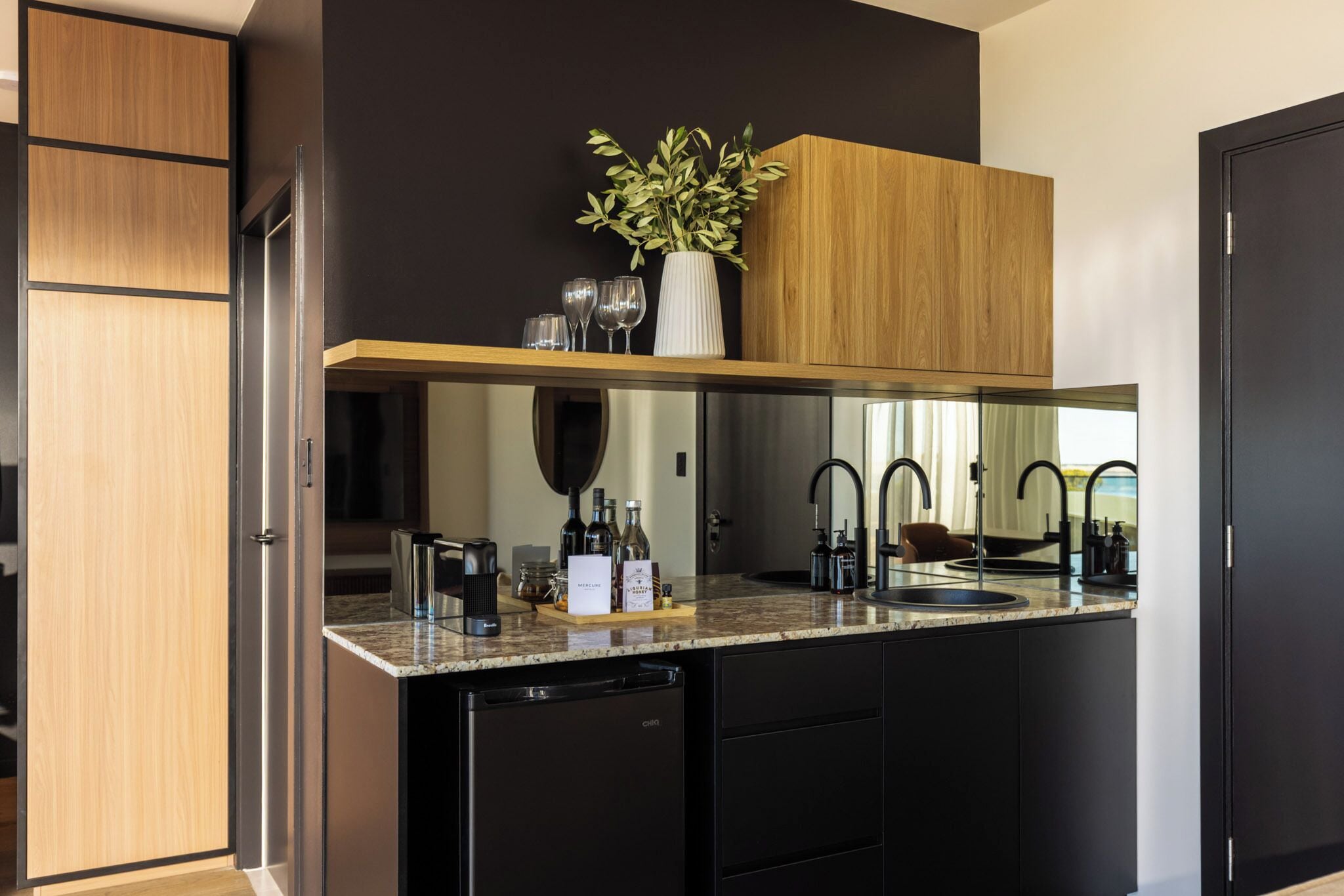 A modern kitchen with a built-in dishwasher, a wooden cabinet, a countertop with a vase of green leaves, and a mirror above the sink.