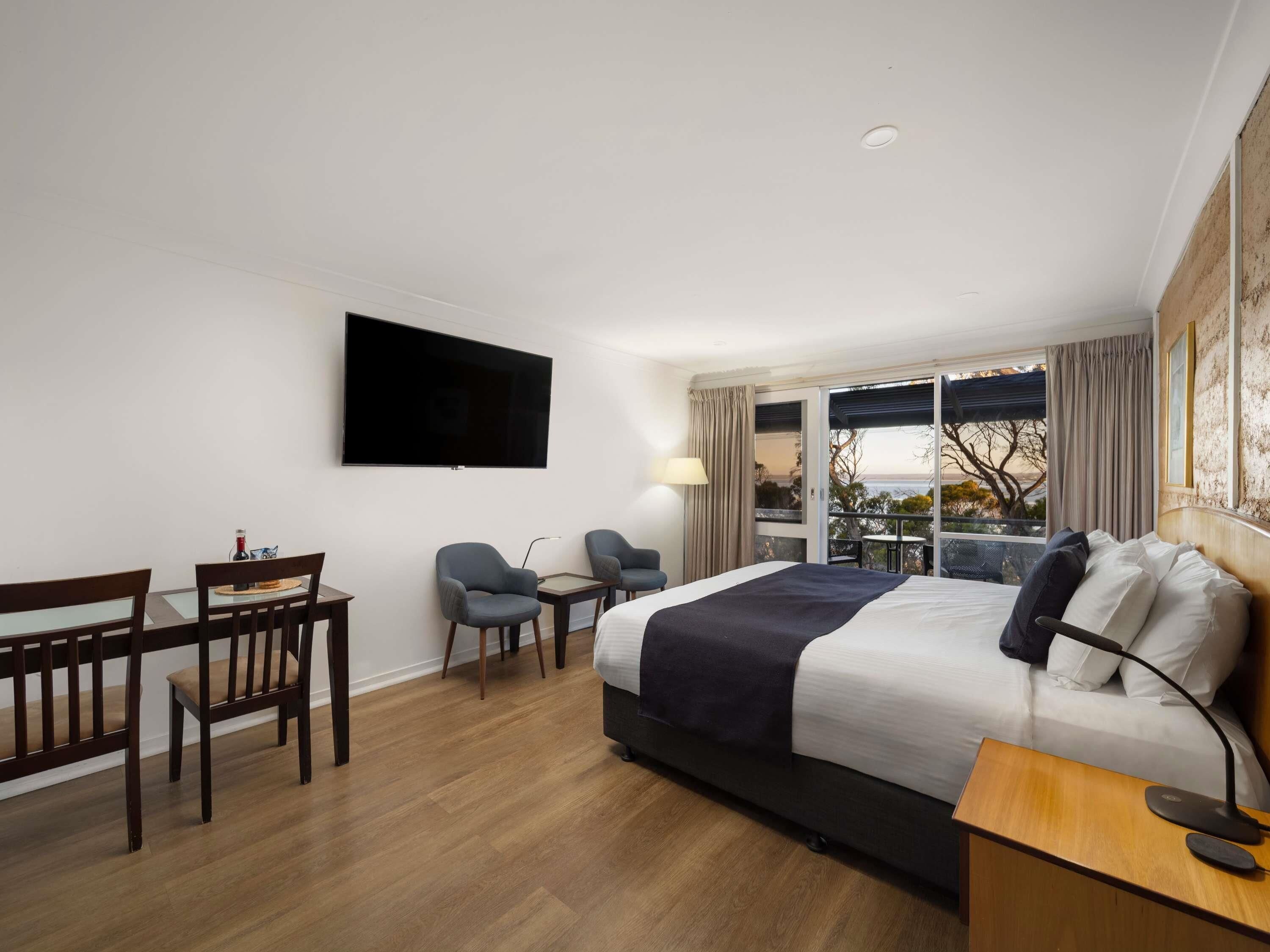 Quarto de hotel com cama, escrivaninha, duas cadeiras, televisão e varanda com vista.