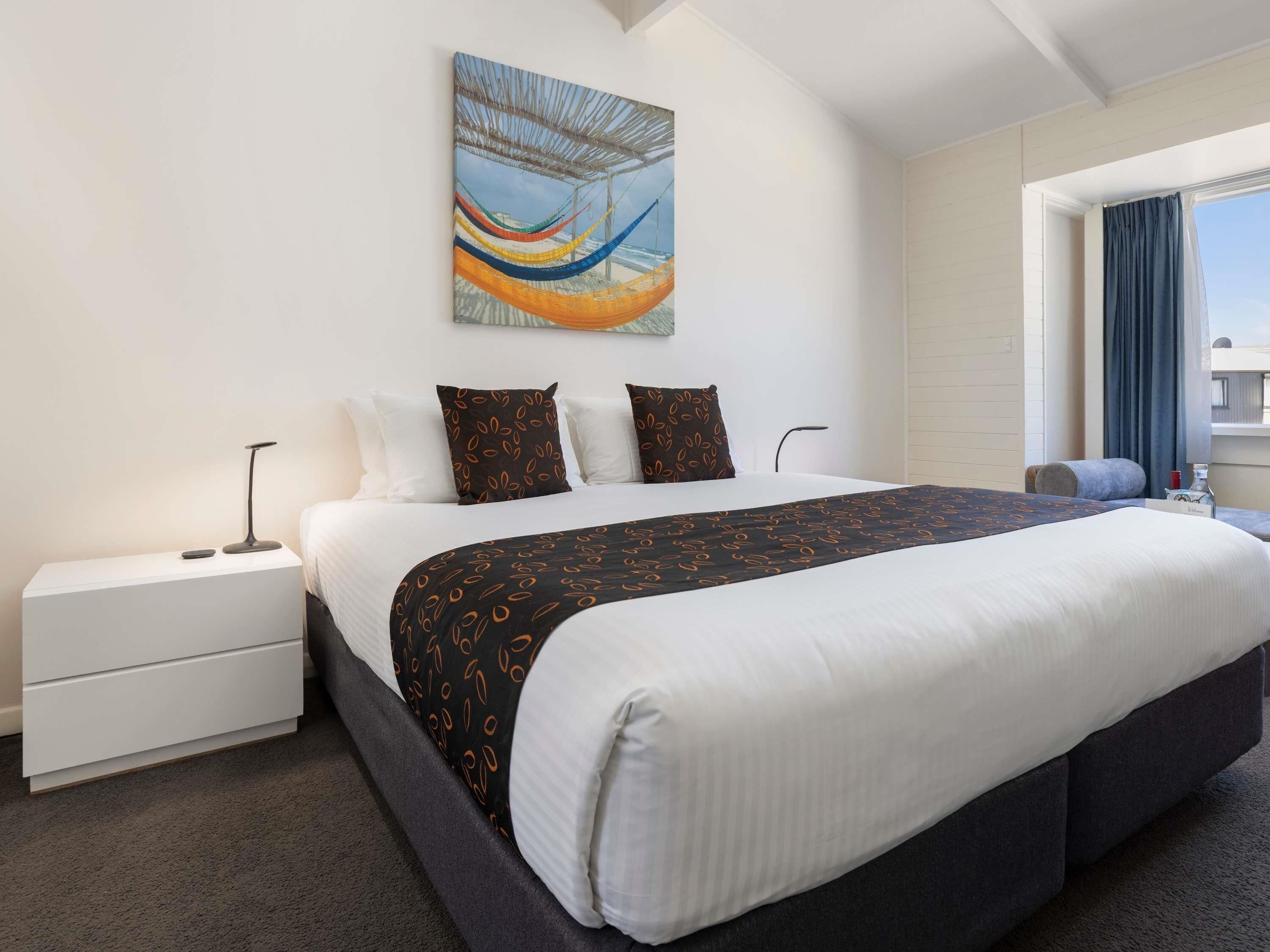 Een hotelkamer met een groot bed, een nachtkastje, een lamp, een schilderij, een raam met gordijnen en uitzicht op de lucht.