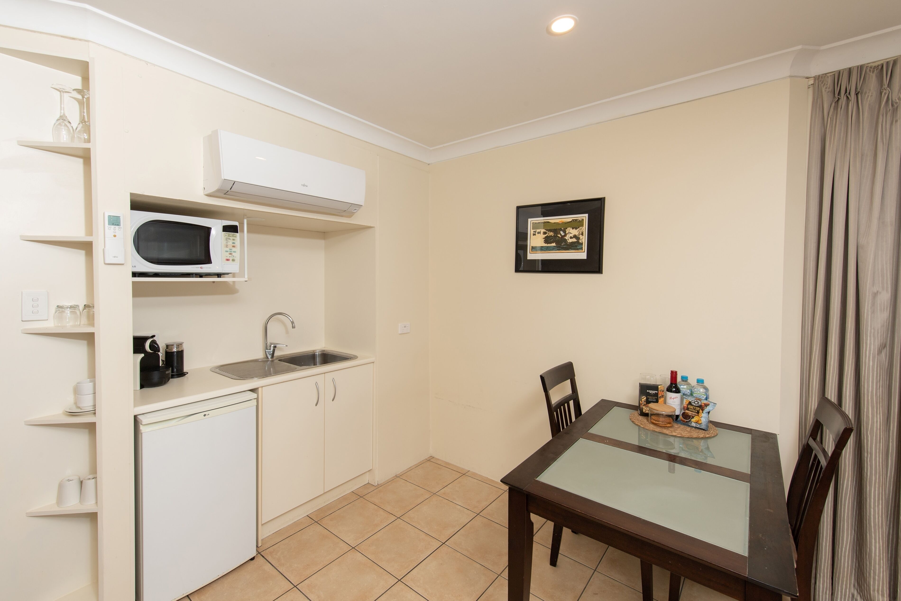 A compact kitchen with a microwave, sink, and a small refrigerator. There is a dining table with chairs and a framed picture on the wall.
