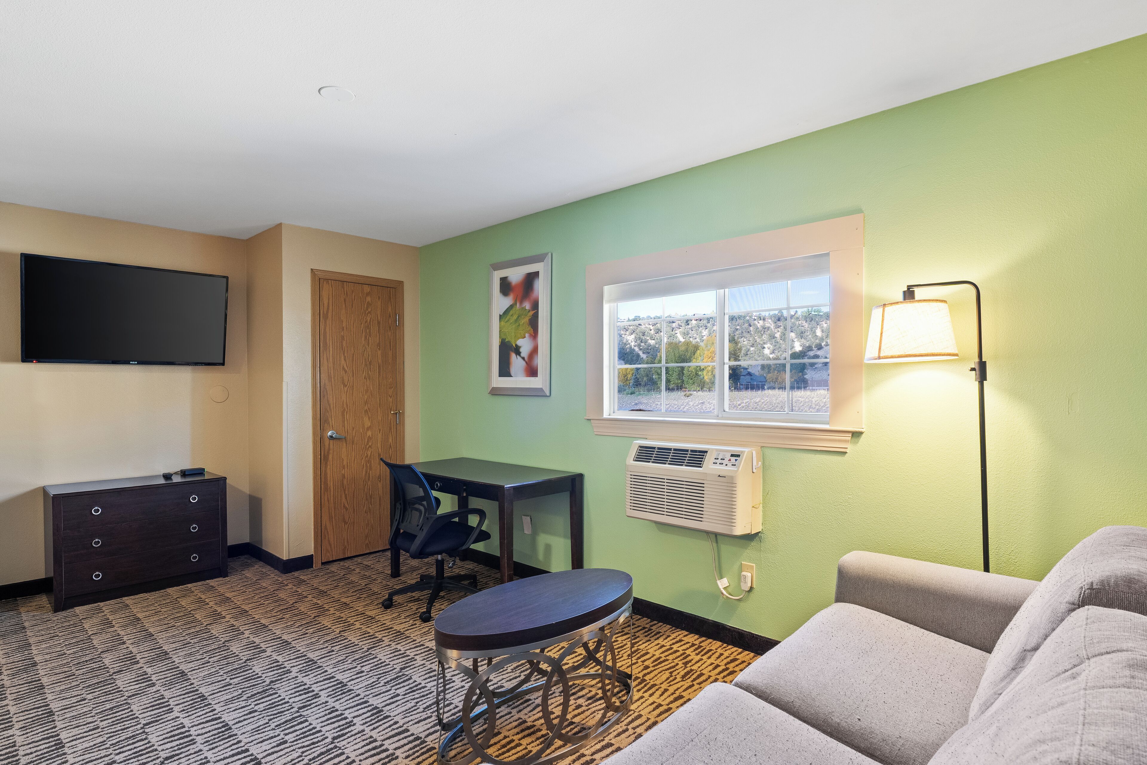 A room with a flat-screen TV, a desk with a chair, a couch, a window with a view, and a wall-mounted air conditioner.