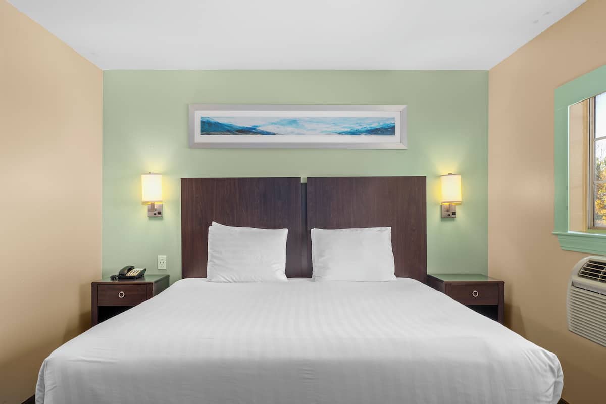 Quarto de hotel com uma cama grande, duas mesas de cabeceira, um criado-mudo, um quadro emoldurado na parede e luminárias fixadas na parede.