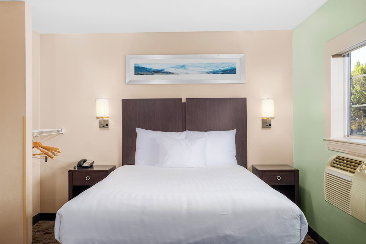 Quarto de hotel com uma cama grande, duas mesas de cabeceira, um criado-mudo, telefone fixado na parede, um quadro sobre a cama e uma janela com unidade de ar condicionado.