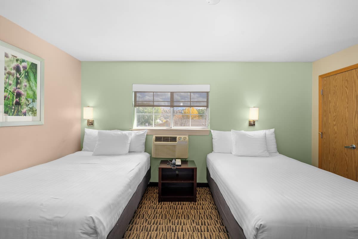 Quarto de hotel com duas camas, janela com vista, mesa de cabeceira com abajur e porta de madeira.