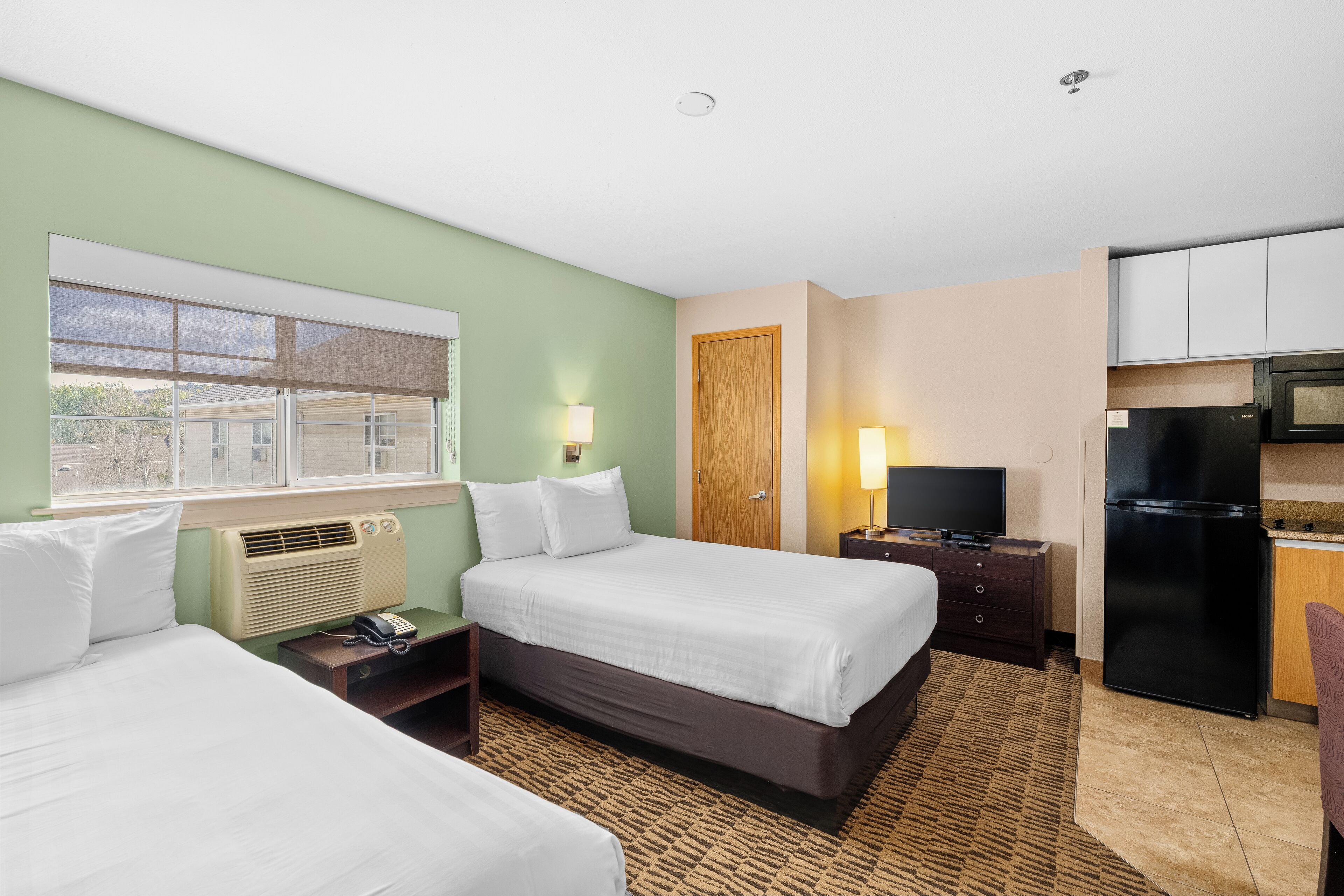 A hotel room with two beds, a kitchenette, a TV, and a window with blinds.