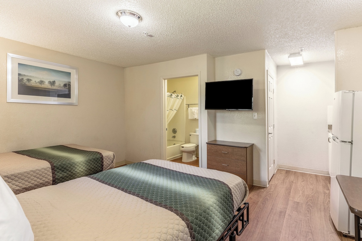 A hotel room with two beds, a television, a bathroom visible through an open door, and a refrigerator.