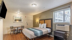 A hotel room with a bed, a desk, a chair, a television, a window, and an air conditioning unit. - HomeTowne Studios by Red Roof Orlando - Casselberry (Casselberry)
