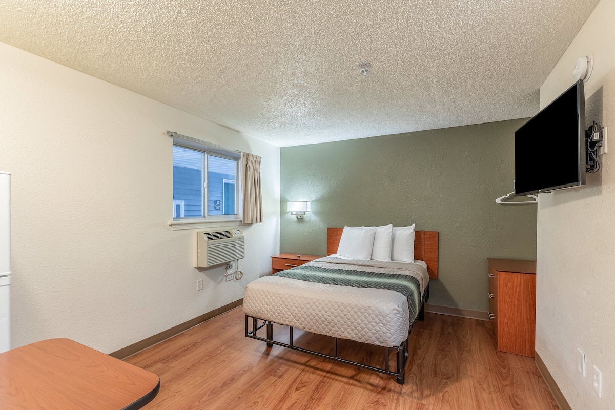 Une chambre d’hôtel comprenant un lit, une table de chevet, une petite armoire en bois, une fenêtre avec des rideaux, un téléviseur fixé au mur et une bouche d’aération au plafond.