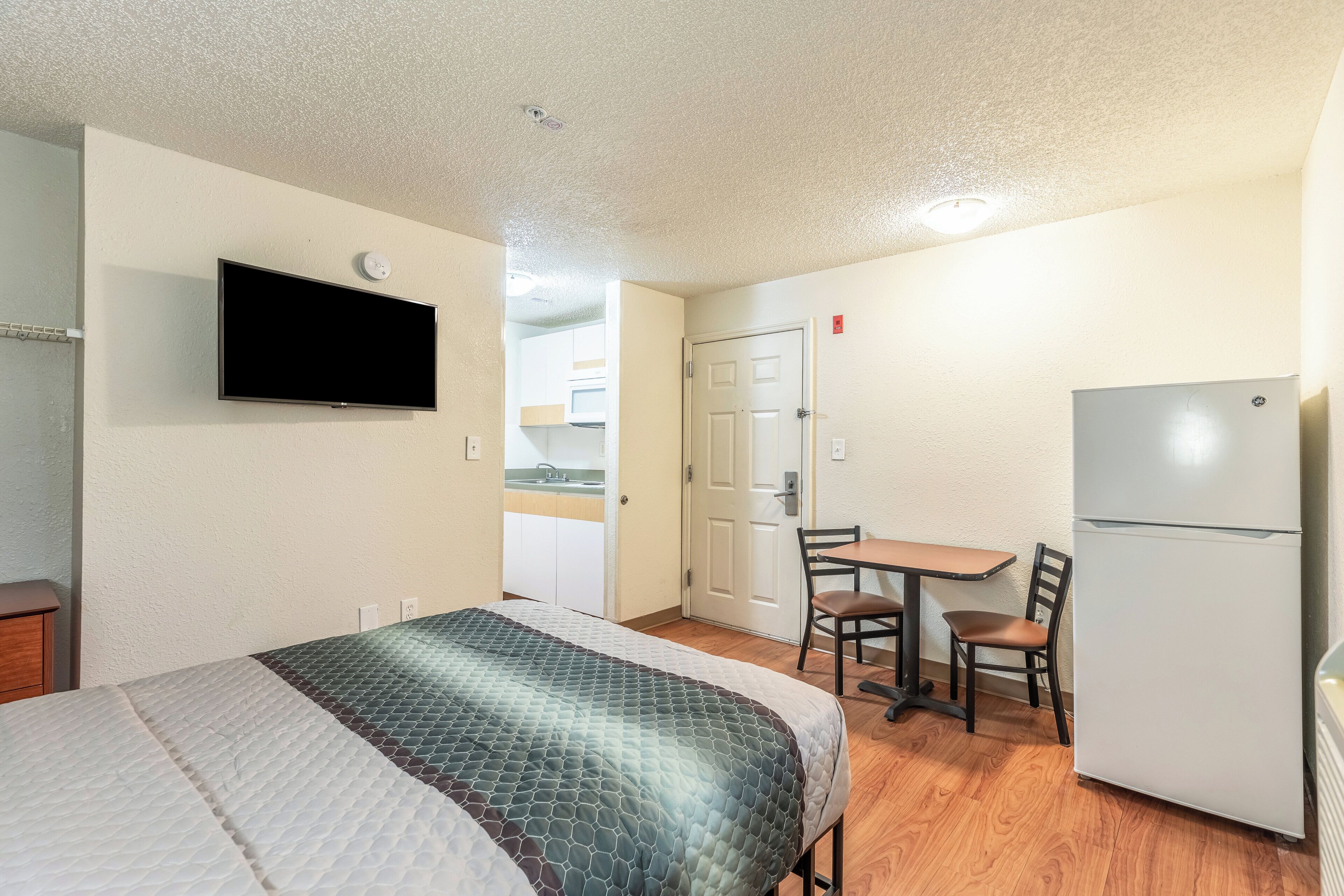 Quarto de hotel compacto com cama, televisão na parede, cozinha compacta e área de refeições com mesa e cadeiras.