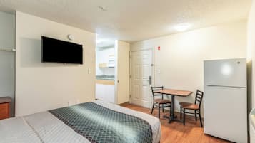 Quarto de hotel compacto com cama, televisão na parede, cozinha compacta e área de refeições com mesa e cadeiras.