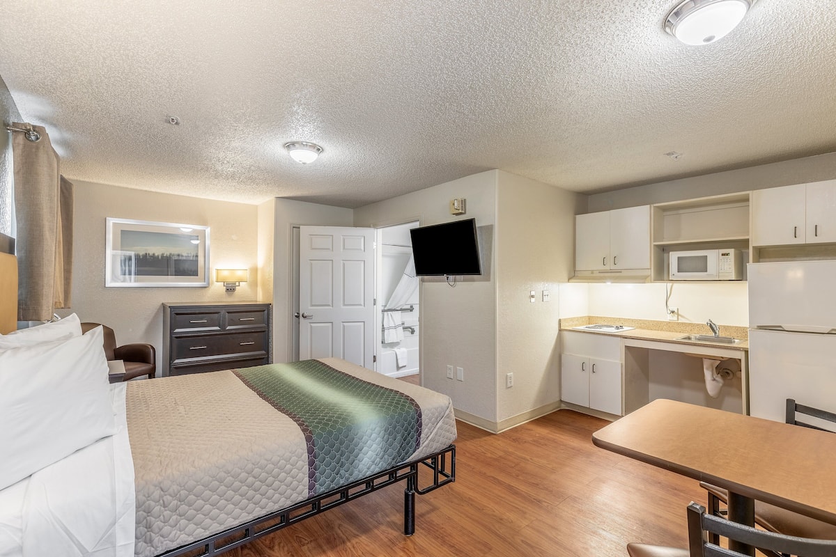 A compact room with a bed, a desk, a chair, a television, a microwave, and a sink.