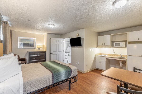 A compact room with a bed, a desk, a chair, a television, a microwave, and a sink.
