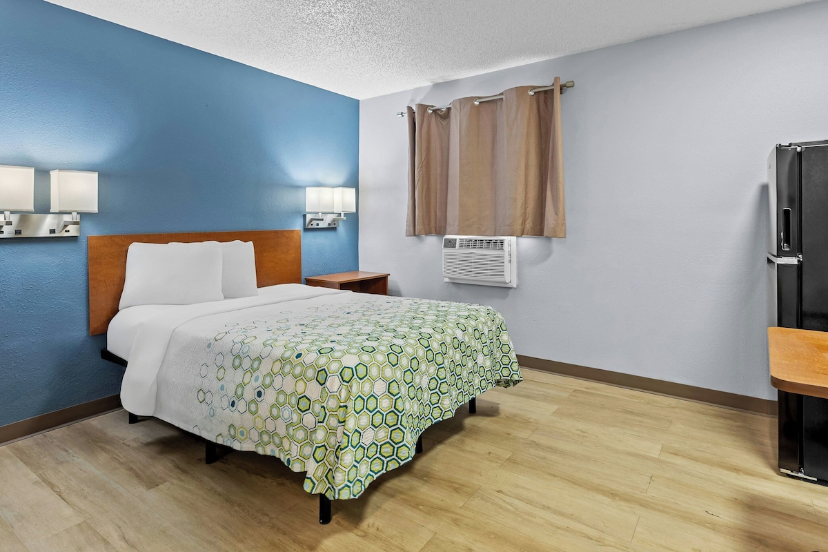 Una habitación de hotel con cama, lámparas de noche, aire acondicionado, refrigerador y ventana con cortinas.