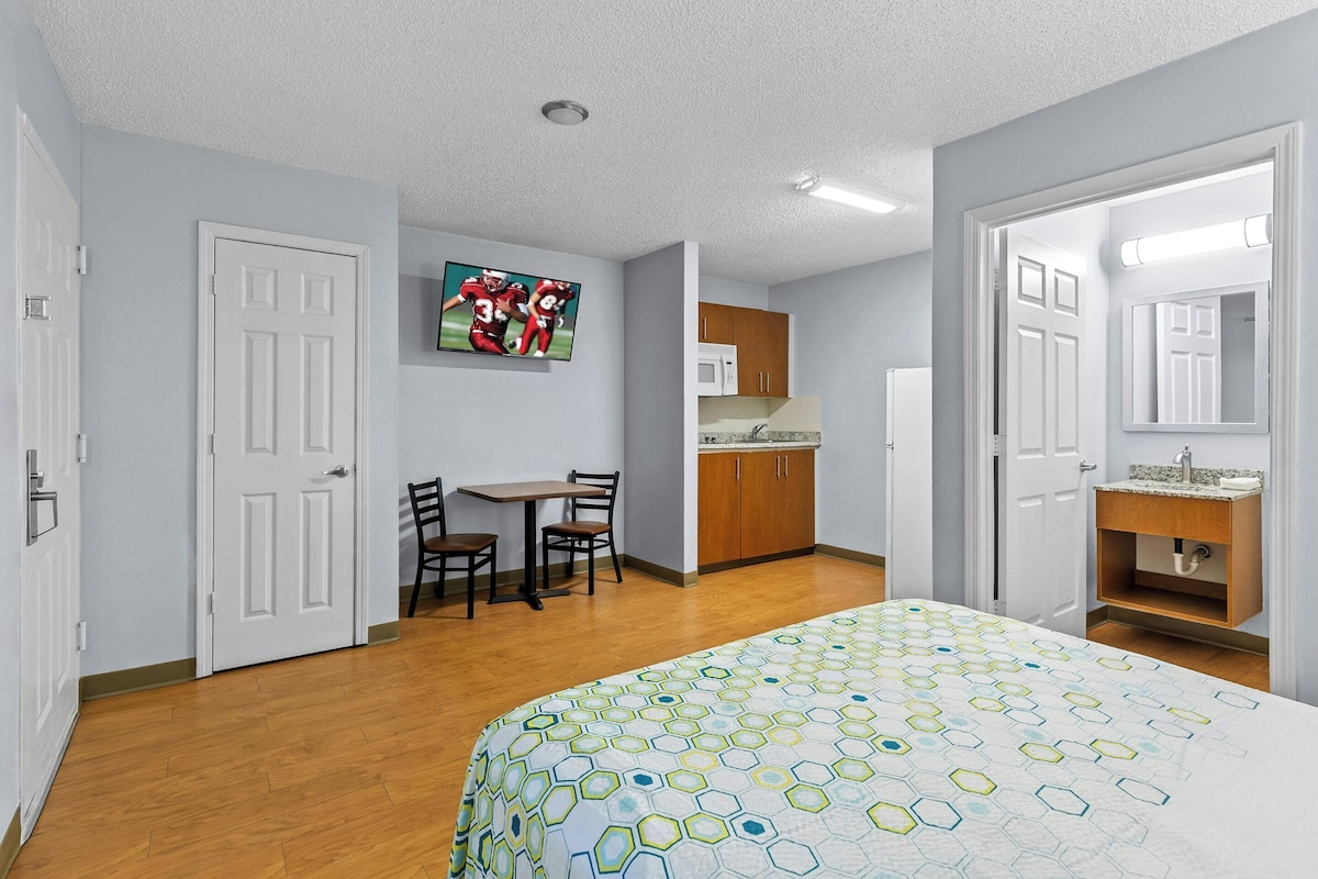 Un dormitorio con cama, escritorio, sillas, televisor, zona de cocina y baño.